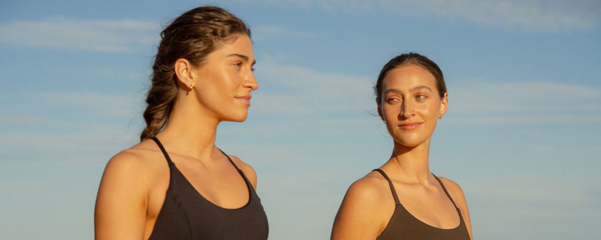 Two women standing side by side in activewear, looking at each other against a blue sky background.